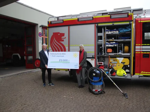 Zwei Menschen stehen vor einem Feuerwehrwagen und halten einen überdimensionierten Scheck in den Händen.
